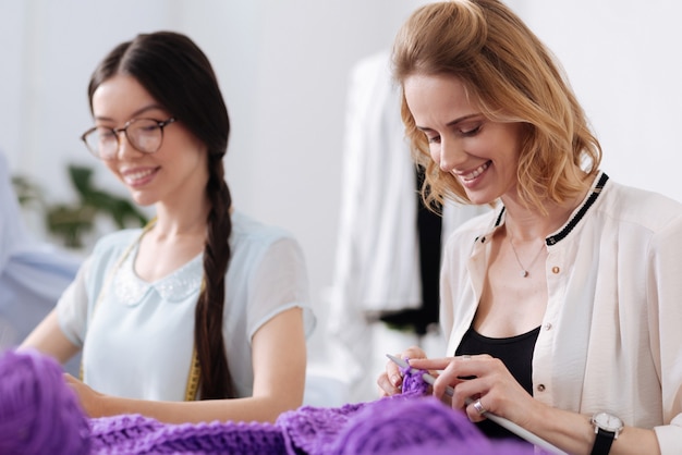 Foto dos hermosas mujeres agradables tejiendo con hilos morados y aparentemente divirtiéndose durante este pasatiempo.