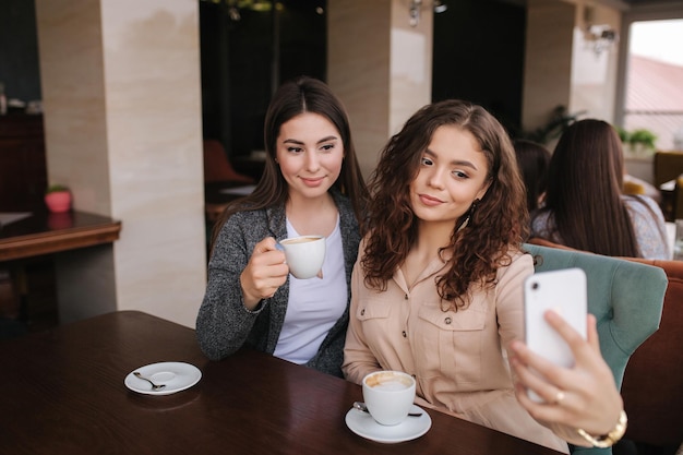 Dos hermosas mujeres hacen selfie en café Mujer de pelo rizado bebe café