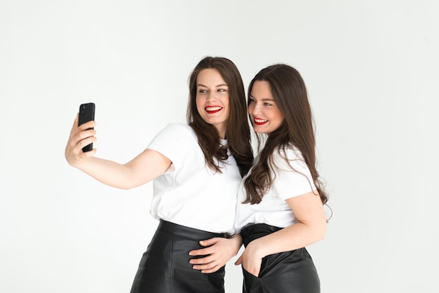 Dos hermosas mujeres hermanas gemelas divirtiéndose juntas posando fondo blanco y tasing selfie