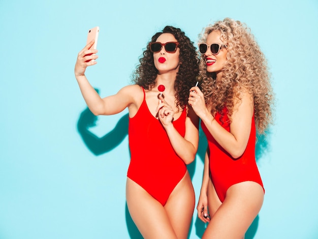 Dos hermosas mujeres hipster sonrientes en trajes de baño rojos Modelos de moda con peinado de rizos tomando selfie en el estudio Mujer caliente posando cerca de la pared azul en gafas de sol Sosteniendo piruleta