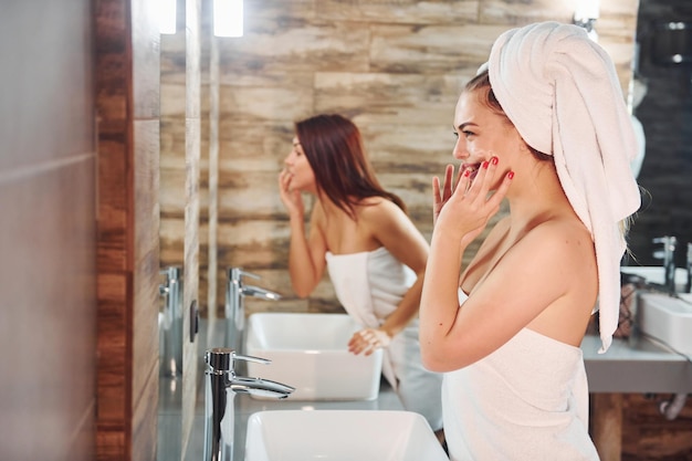 Dos hermosas mujeres jóvenes juntas en el baño cerca del espejo y limpiándose la cara.