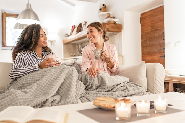 Dos hermosas mujeres jóvenes sentados en el sofá con una manta en las piernas riendo disfrutando de la casa en invierno tomando té con pasteles - Estilo de vida en casa de pareja femenina de raza mixta