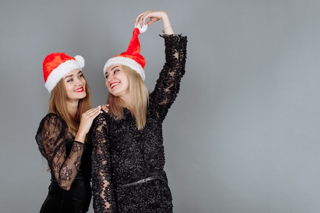 Dos hermosas mujeres jóvenes con vestidos negros y gorros de santa divirtiéndose y celebrando una fiesta de Navidad en el estudio.