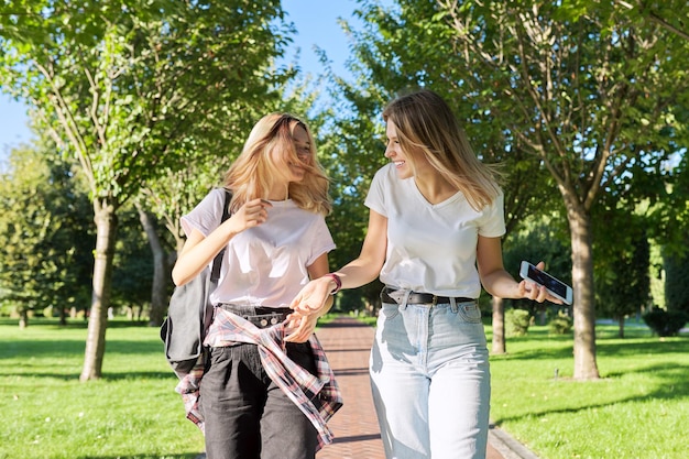 Dos hermosas niñas adolescentes felices de 17, 18 años caminando juntos en el parque, niñas riendo hablando divirtiéndose en un día soleado de verano