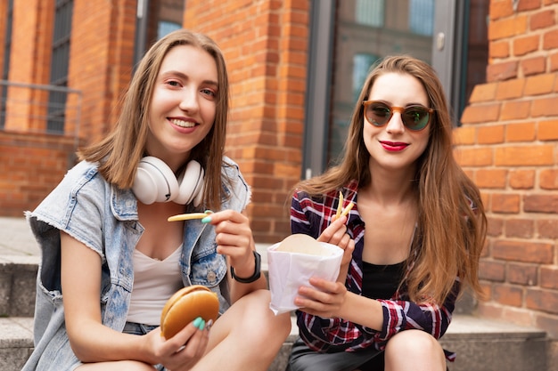 Dos hermosas niñas comen comida rápida en la calle, se divierten, hablan y se alimentan mutuamente cosas deliciosas.