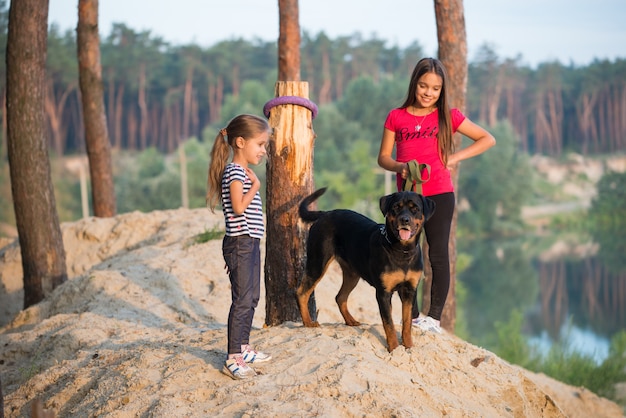 Dos hermosas niñas lindas y un gran perro Rottweiler adulto, sentados en el borde de un acantilado de arena abrazándose y disfrutando de una cálida mañana de verano en un denso bosque de abetos