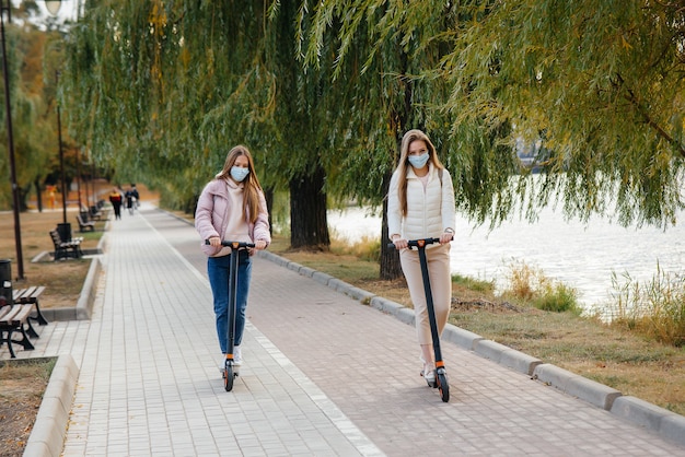 Dos hermosas niñas con máscaras andan en patinetes eléctricos en el parque en un cálido día de otoño. Caminar en el parque.