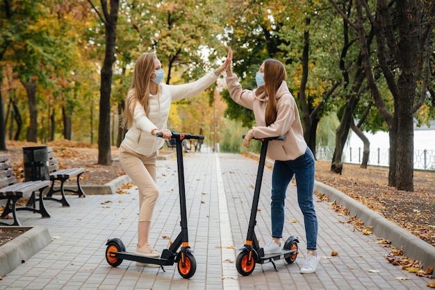 Dos hermosas niñas con máscaras andan en patinetes eléctricos en el parque en un cálido día de otoño. Caminar en el parque.