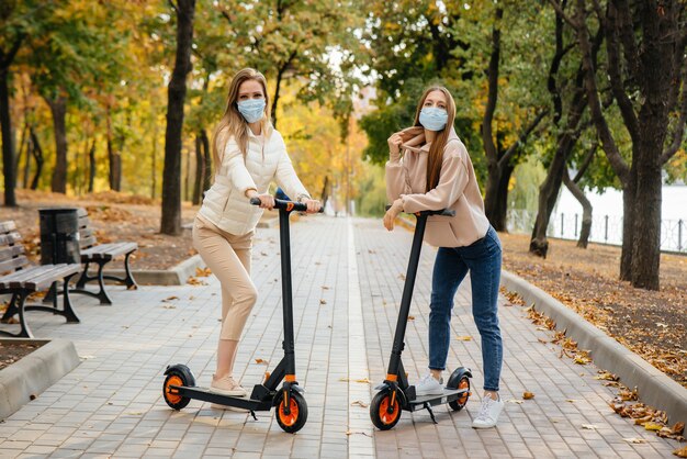 Dos hermosas niñas con máscaras andan en patinetes eléctricos en el parque en un cálido día de otoño. Caminar en el parque.