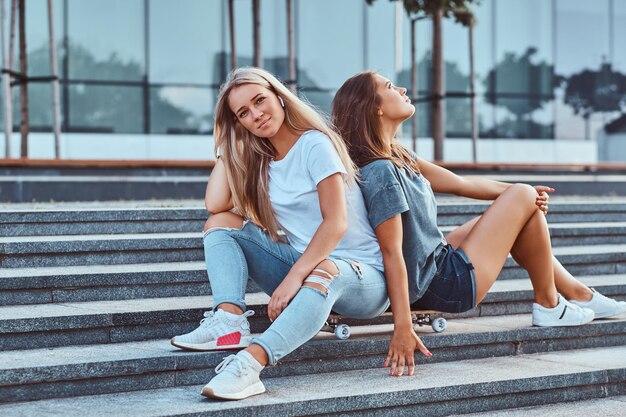 Dos hermosas novias jóvenes sentadas en una patineta en pasos sobre un fondo del rascacielos.