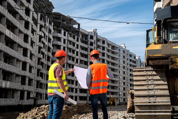 Dos hermosos trabajadores en uniforme en un sitio de construcción están considerando un plan. Equipos de construcción