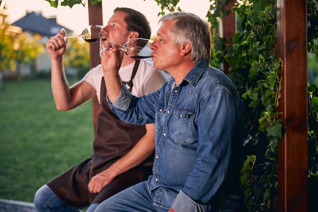 Dos hombres adultos sentados en el jardín con vasos de vino tinto y tomando un sorbo simultáneamente