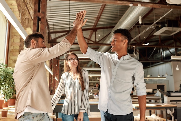 Dos hombres alegres dando highfive mientras está de pie con sus colegas en el trabajo en equipo de la oficina moderna