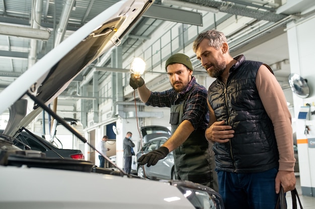 Dos hombres barbudos inclinándose sobre el compartimiento del motor del automóvil abierto mientras el técnico con la lámpara apuntando a una de sus partes durante el diagnóstico