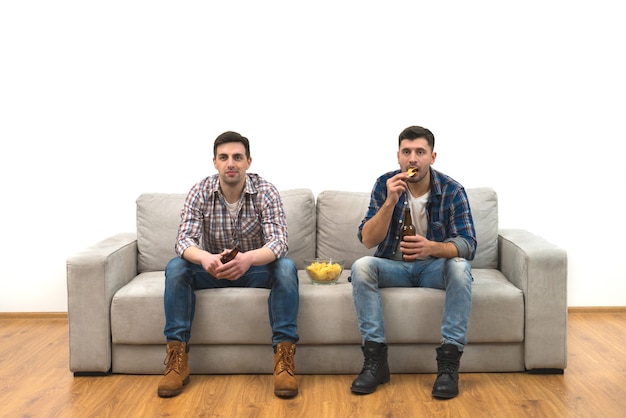 Los dos hombres beben una cerveza en el sofá sobre una pared blanca de fondo