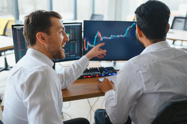 Dos hombres comerciantes sentados en el escritorio en la oficina juntos monitoreando gráficos de velas de datos de acciones en la pantalla analizando el flujo de precios sonriendo alegremente teniendo un concepto de trabajo en equipo de ganancias