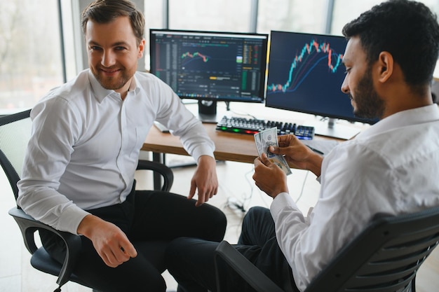 Dos hombres comerciantes sentados en el escritorio en la oficina juntos monitoreando gráficos de velas de datos de acciones en la pantalla analizando el flujo de precios sonriendo alegremente teniendo un concepto de trabajo en equipo de ganancias