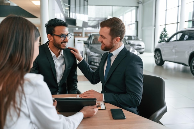 Dos hombres están comprando un automóvil tres personas están trabajando juntas en la sala de exposición de automóviles