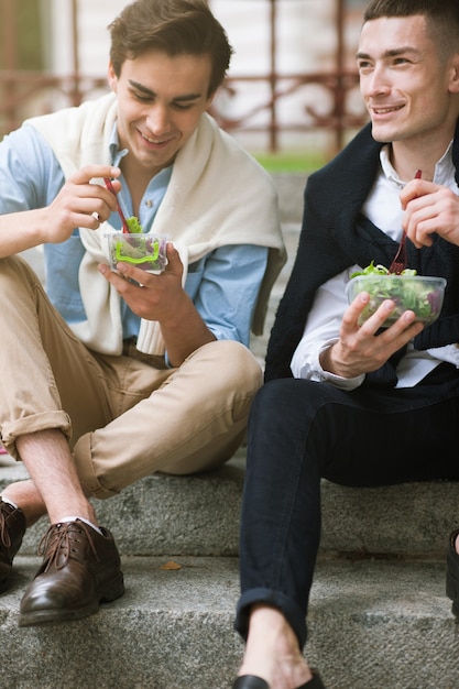Dos hombres felices con comida sana al aire libre. Estilo callejero masculino. Pausa para el almuerzo, jóvenes alegres con estilo, concepto de dieta
