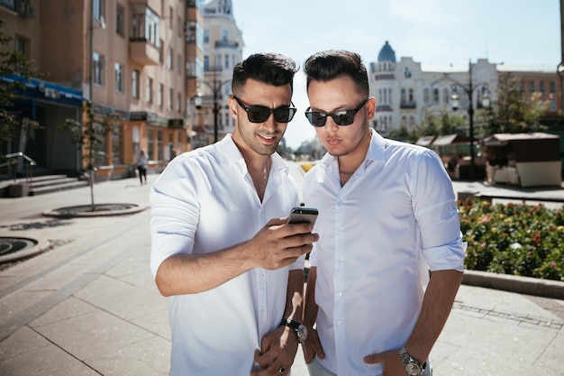 Dos hombres felices riendo y mirando el teléfono en la calle en el día soleado y discutiendo algo interesante