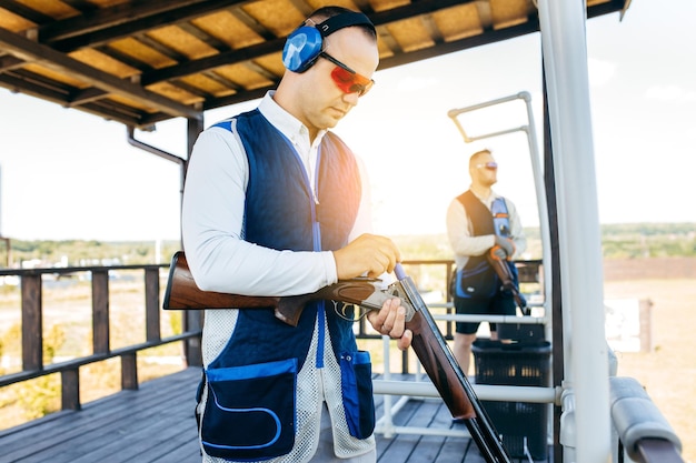 Dos hombres con gafas de sol, auriculares protectores y un chaleco de rifle practicando disparos con armas de fuego