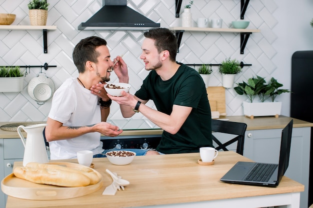 Dos hombres gay jóvenes felices mirando el uno al otro con una sonrisa durante el desayuno. Hombre alimentando a su pareja