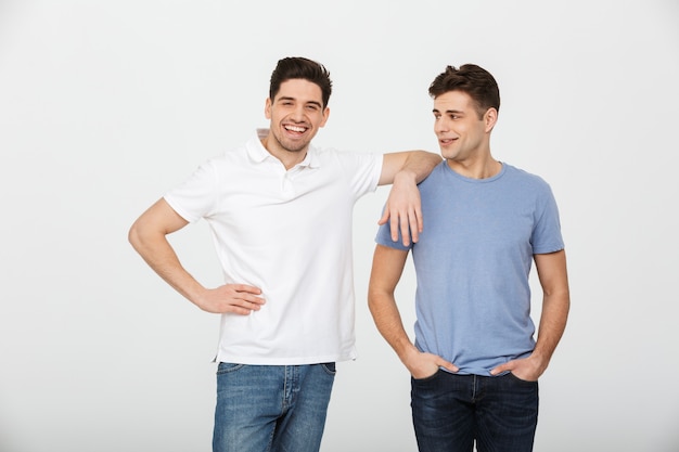 Dos hombres guapos muchachos 30s vistiendo camiseta casual y jeans sonriendo y posando juntos en la cámara, aislado sobre fondo blanco.