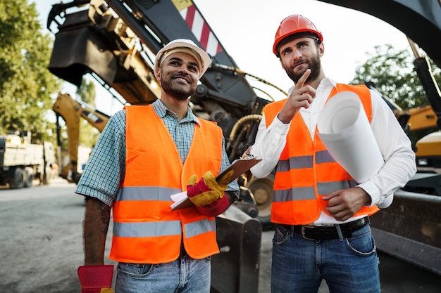 Dos hombres ingenieros discutiendo su trabajo de pie contra las máquinas de construcción