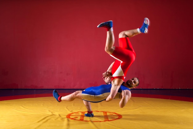 Dos hombres jóvenes en mallas de lucha azules y rojas están luchando y haciendo una lucha suplex en una alfombra de lucha libre amarilla en el gimnasio