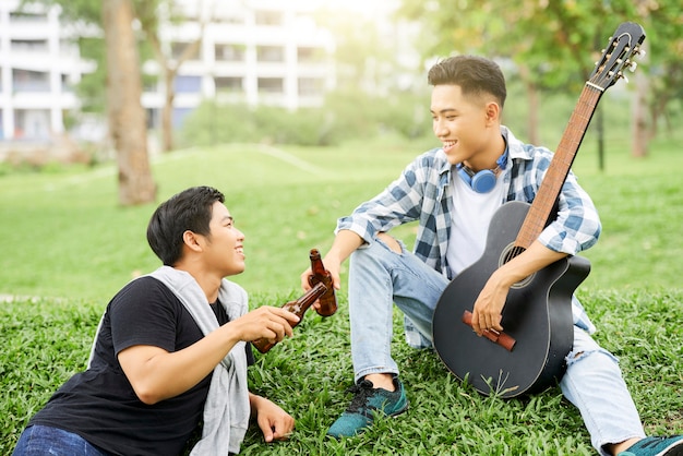 Dos hombres jóvenes sentados sobre la hierba verde con guitarra y bebiendo cerveza al aire libre