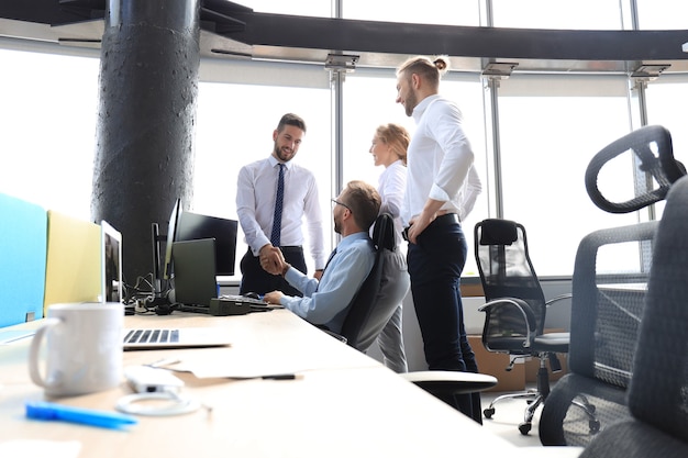 Dos hombres de negocios alegres estrecharme la mano mientras están en la oficina junto con sus colegas.