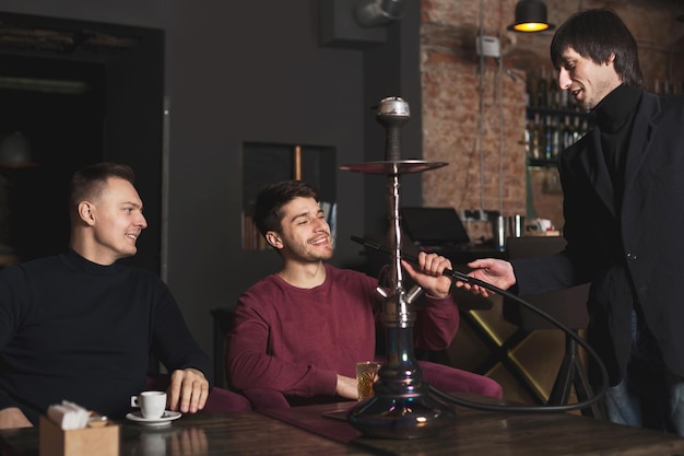 Dos hombres de negocios confiados bebiendo whisky y pidiendo narguile, teniendo una reunión de negocios informal en el café, espacio de copia