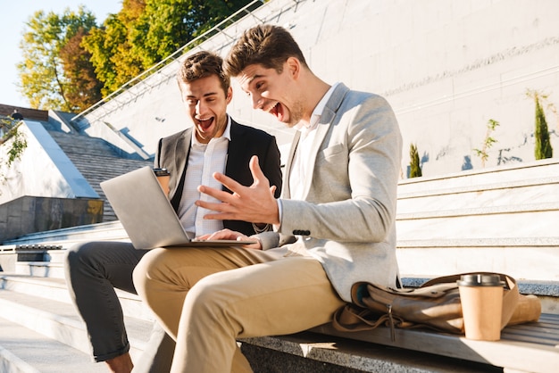 Dos hombres de negocios confiados usando la computadora portátil mientras están sentados en un banco al aire libre, celebrando el éxito