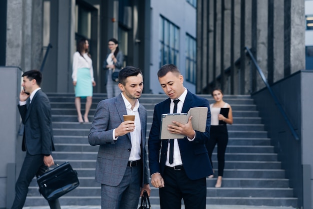 Dos hombres de negocios guapos caucásicos en trajes y corbatas con café en las manos y caminando desde el edificio de oficinas después del día de trabajo. Hombres con estilo hablando y discutiendo negocios.