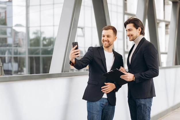 Dos hombres de negocios modernos tomándose selfies o haciendo una videollamada en el contexto de oficinas y edificios urbanos