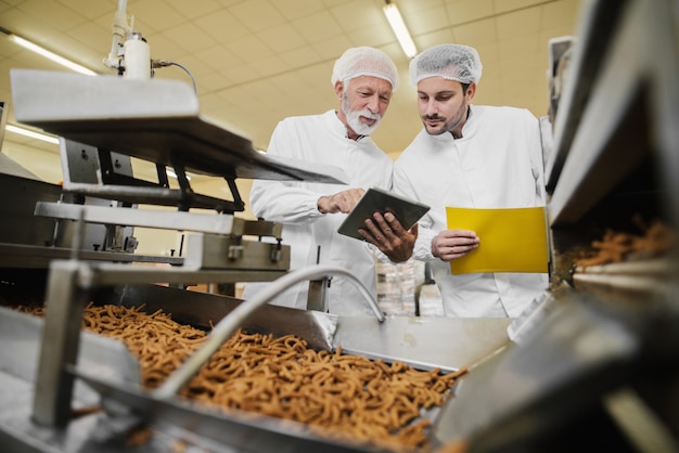 Dos hombres de negocios en ropa estéril de pie en la fábrica de alimentos frente a la línea de producción y mirando y tableta. Comprobando la calidad de los productos y hablando.