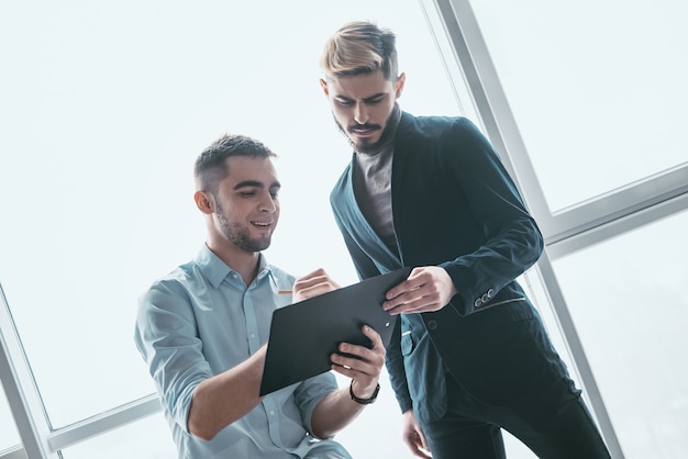 Dos hombres de negocios sonrientes hablando de inversión elegible, gerente presentando un informe financiero que muestra buenos resultados de trabajo a su colega, señalando en papel, ofreciendo trato, discutiendo un nuevo proyecto