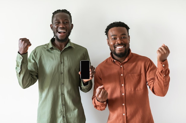 Dos hombres negros alegres que muestran la pantalla del teléfono inteligente sobre fondo blanco