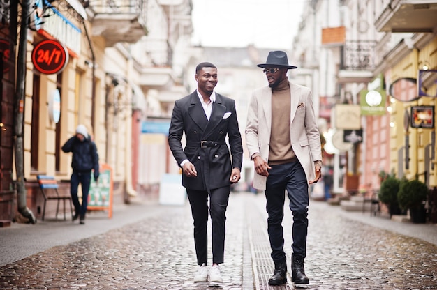 Dos hombres negros de moda caminando en la calle. Retrato de moda de modelos masculinos afroamericanos. Use traje, abrigo y sombrero.