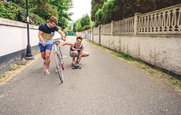Dos hombres riendo y divirtiéndose montando bicicleta y patineta en la calle en un día soleado. Concepto de estilo de vida joven.