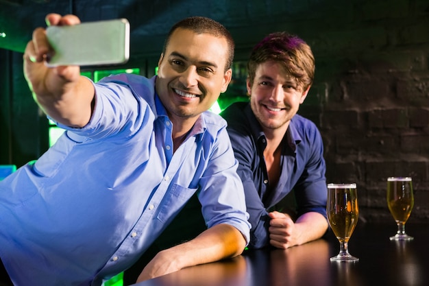 Dos hombres tomando una selfie en el teléfono mientras toman cerveza en la barra de bar en el bar