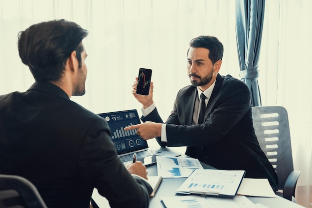 Dos hombres con traje se sientan en un escritorio, uno de ellos sostiene un teléfono y el otro mira una pantalla que dice "plan de negocios".
