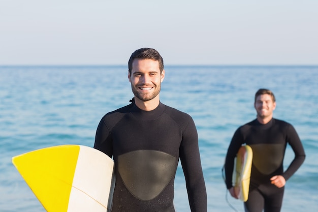 Dos hombres en trajes de neopreno con una tabla de surf en un día soleado