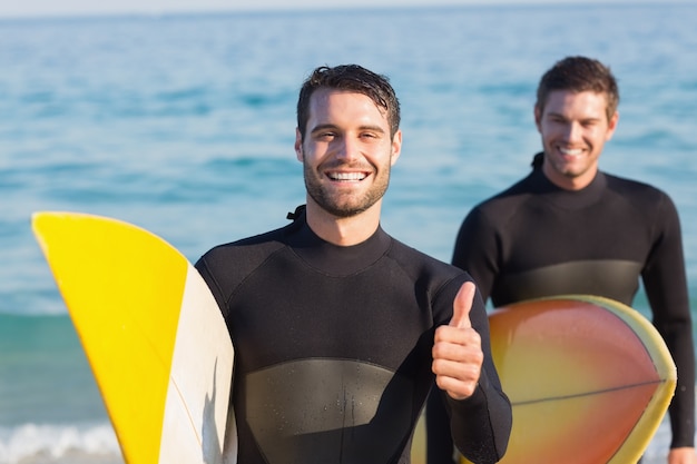 Dos hombres en trajes de neopreno con una tabla de surf en un día soleado