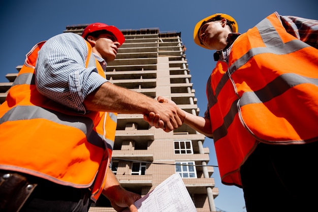 Dos hombres vestidos con camisas, chalecos de trabajo naranja y cascos se dan la mano en el contexto de un edificio de varios pisos.