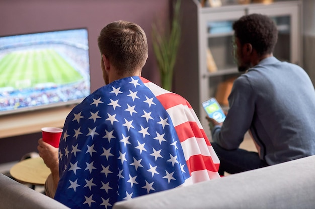 Dos hombres viendo un partido de fútbol en la televisión y usando una bandera estadounidense