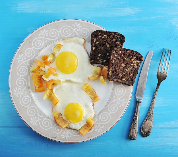 Dos huevos fritos con pimienta y dos rebanadas de pan de centeno en un plato, cuchillo y tenedor sobre una superficie de madera azul