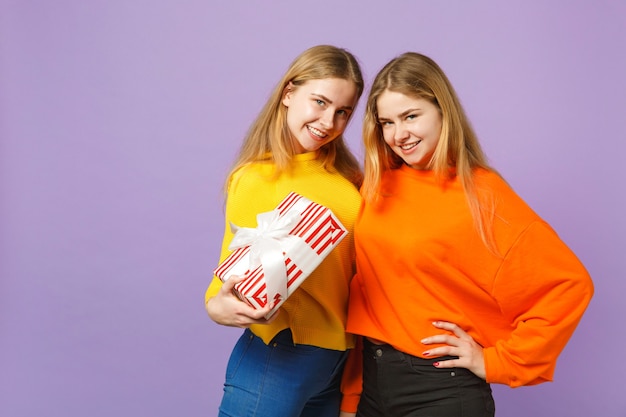 Dos impresionantes hermanas gemelas rubias niñas en ropa vívida sostienen la caja presente de rayas rojas con cinta de regalo aislada en la pared azul violeta. Cumpleaños de la familia de personas, concepto de vacaciones.