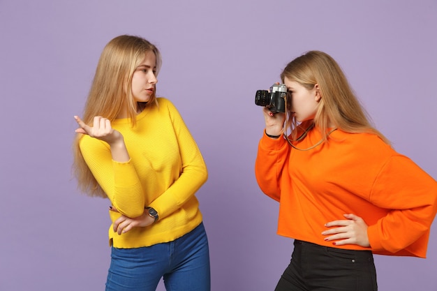 Dos impresionantes niñas hermanas gemelas rubias jóvenes en ropa colorida tomando fotografías en la cámara de fotos vintage retro aislada en la pared azul violeta. Concepto de estilo de vida familiar de personas.