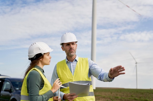 Dos ingenieros caucásicos de pie en el campo de la turbina eólica y discutiendo sobre documentos